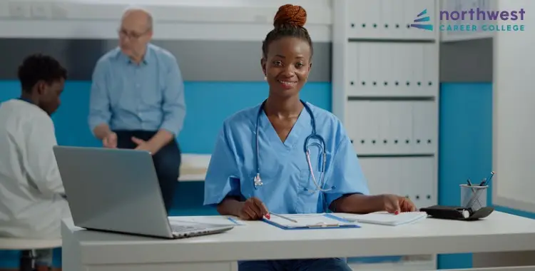 A smiling Medical Assistant in scrubs sits at a desk with a laptop, while patients are seen in the background.