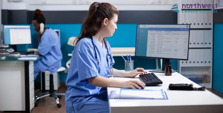 A Medical Administrative Assistant works at a computer in a healthcare setting, managing patient information.