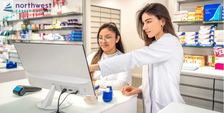 Two Pharmacy Technicians collaborate at a computer in a bright pharmacy, surrounded by shelves of medications.