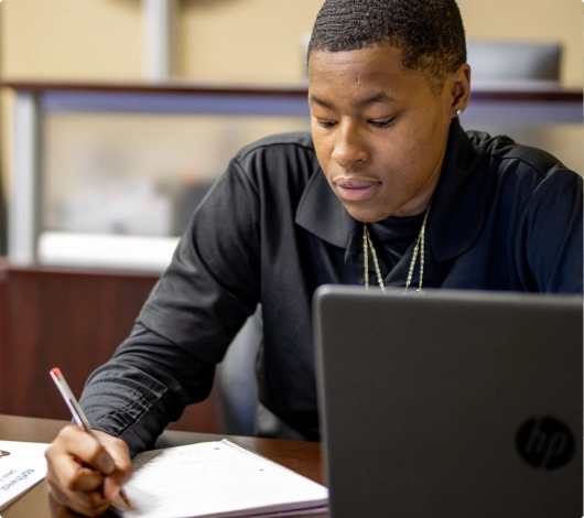 A student engages with coursework in a medical administrative assistant program, writing notes beside a laptop.