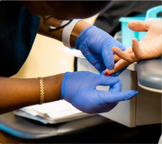 A phlebotomy technician program student draws blood from a patients finger while wearing blue gloves.