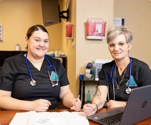 Two certified medical assistants sit at a table, smiling, with a laptop and medical supplies in a clinical setting.