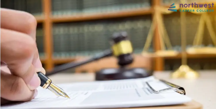 A hand holding a pen writes on a clipboard, with a gavel and legal scales in the background, suggesting Legal Assistant.