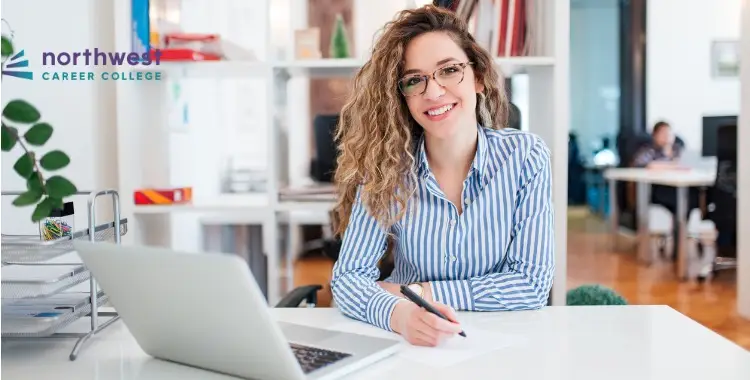 A smiling woman with curly hair and glasses sits at a desk, representing Business Administrative Assistants at Northwest.