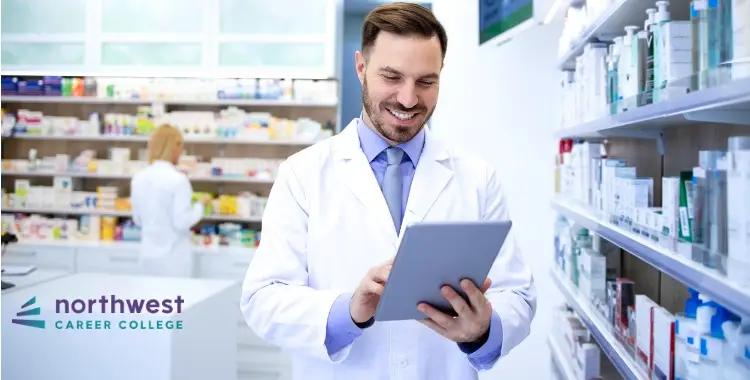 A Pharmacy Technician smiles while using a tablet in a well-stocked pharmacy, with another technician in the background.