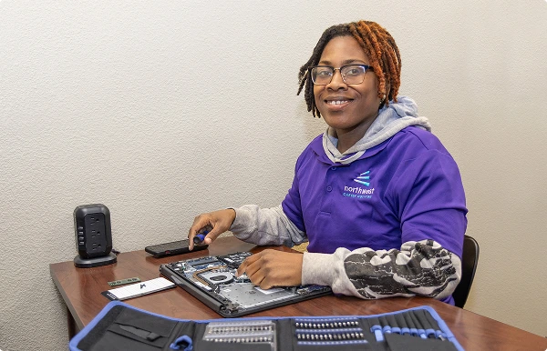 A student at an IT technician trade school works on a laptop, smiling with tools and a phone nearby.