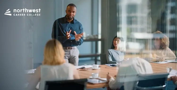 A man presents to a group in a meeting, discussing topics from the Guide to Business Administration.