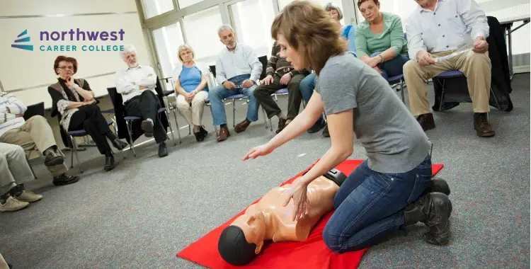 A woman performs CPR on a mannequin while an audience watches in a classroom setting.