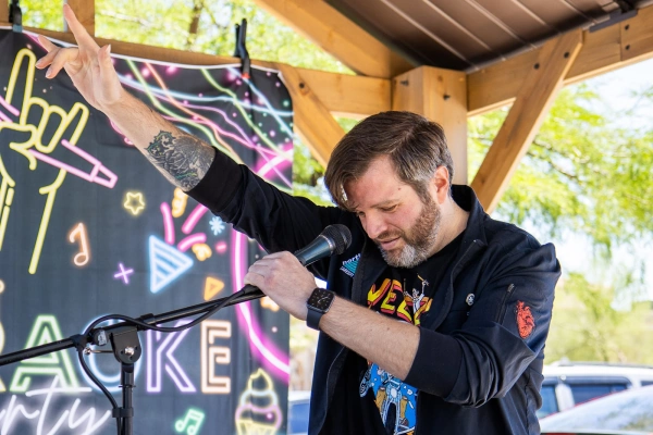 A man with a beard sings into a microphone, raising one arm, in front of a colorful backdrop at an outdoor event.